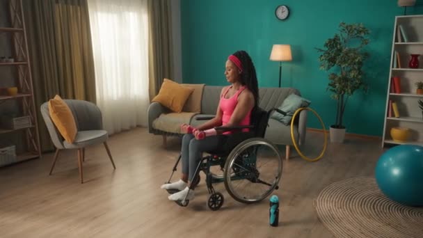 リビングルームでダンベルで運動する車椅子のアフリカ系アメリカ人女性 ピンクのヘッドバンドとトラックスーツの女性は 腕の筋肉を訓練し 彼女の足とトリップを振る — ストック動画
