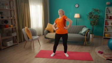 Gözlüklü yaşlı bir kadın ellerini beline koyar ve vücut dönüş egzersizi yapar. Emekli bir kadın spor için evine gidiyor. Yaşlılar için aktif yaşam tarzı ve emeklilik kavramı