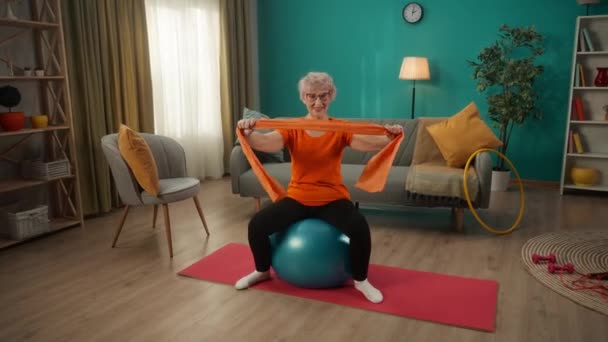 一位老年妇女坐在一个适配球上 带着一个运动弹性带在客厅里训练 一个女人手里拿着一个膨胀器 把胳膊伸向两边 增强了肌肉 — 图库视频影像