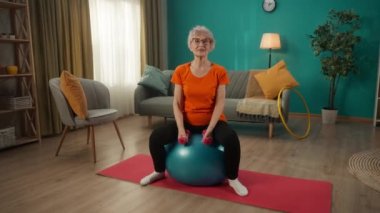 Yaşlı bir kadın bir fitbol topuna oturur ve oturma odasında dambıllarla antrenman yapar. Kadın kol ve kol kasları için egzersiz yapıyor. Vücudunu formda tut. Spor yaşam tarzı, emeklilik, sağlık kavramı