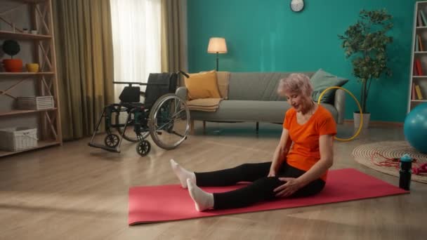 一名行动不便的老年妇女坐在轮椅旁边客厅的地板上 一个灰白头发的女人给自己按摩了一下脚 身体康复的概念 — 图库视频影像