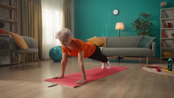 一位穿着运动服的老年妇女躺在电话前的地板上 一个女人开始在她的电话计时器和进入一个木板的位置 家庭健身概念 在线体育课 — 图库视频影像