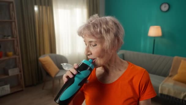 stock video Portrait of an exhausted but satisfied grayhaired elderly woman after a workout in the living room. Tired elderly woman drinks water after exercise. The concept of sports lifestyle, retirement