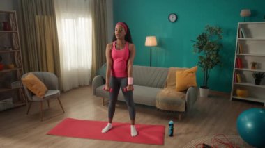 Genç atletik kadın spor için evine gidiyor. Bir kadın kollarının ve omuzlarının kaslarını dambıllarla eğitir. Afro-Amerikalı bir kadın kollarını kaldırır ve dambıllarını öne doğru uzatır.