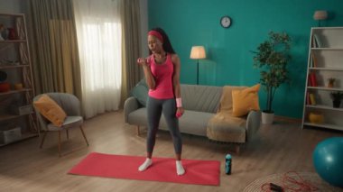 Genç, formda bir kadın evde halterlerle kol egzersizi yapıyor. Afrikalı bir Amerikalı kadın kol kaslarını güçlendirmek için halterleri acıdan ve yorgunluktan kaldırır. Spor konsepti, ev sporu.