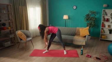 Oturma odasında egzersiz yapmadan önce bir kadın ısınıyor. İnce, atletik Afrikalı Amerikalı bir kadın yan eğilip öne doğru eğiliyor. Ev fitness konsepti. Yavaş çekim