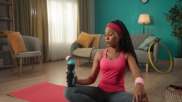 アフリカ系アメリカ人女性は自宅で激しいワークアウトの後に休憩を取る 女性は彼女の額に手を置き 安心して追放し 喜びで水を飲む ボトルを手にした女性が横に座り — ストック動画
