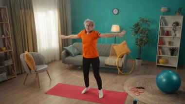 Oturma odasındaki paspasın üzerinde yıldız atlayışı yapan yaşlı bir kadın. Beyaz saçlı, emekli bir kadın sağlığına dikkat eder, bacak ve kalçalarının kaslarını güçlendirir. Sportif yaşam tarzı kavramı