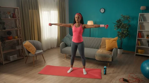 若い運動選手は家でスポーツをする 女性はダンベルで腕と肩の筋肉を訓練します アフリカ系アメリカ人女性がダンベルで腕を横に広げている コンセプト — ストック写真