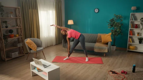 运动的非洲裔美国妇女站在客厅的一个运动垫上做侧弯 一位年轻女子在笔记本电脑上看了一节课录像 然后重复练习 家庭健康概念 — 图库照片