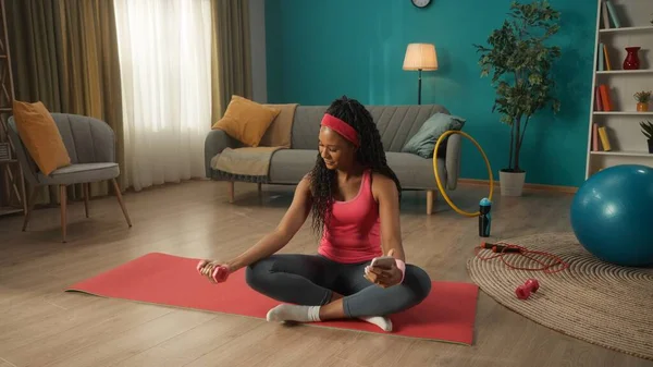 アフリカ系アメリカ人の女性がリビングの床の上のスポーツマットに足を組んで座っている 女性は片手にダンベルを持ち 手を動かすとスマートフォンを持っている — ストック写真