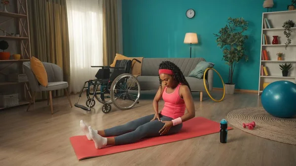 車椅子の横のリビングルームの床には障害者の女性が座っています アフリカ系アメリカ人の若い女性が足のマッサージをしています 身体的リハビリテーションの概念 — ストック写真