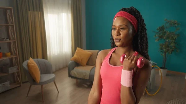 年轻健康的女人在家用哑铃做二头肌运动 一位非裔美国妇女弯下一只胳膊 肘部有一个哑铃 另一只胳膊弯下腰来 家庭健康的概念 — 图库照片