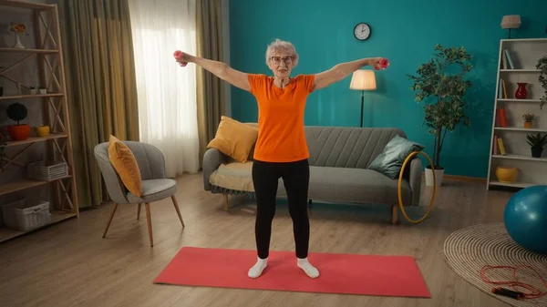 一个退休的女人照顾自己的健康 在家参加体育活动 一个年长的女人带着哑铃训练 锻炼她的手臂肌肉 那个女人用哑铃把胳膊张开 — 图库照片