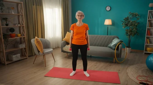 年金受給者は彼女の健康の世話をし 家でスポーツに行く 高齢の女性はダンベルで訓練し 腕の筋肉を強化する 女性がスポーツマットの上に立っていて ダンベルを抱えている — ストック写真
