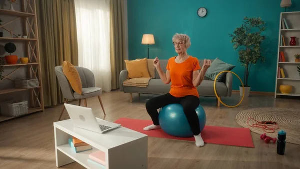 老年妇女坐在笔记本电脑前的健身球上参加体育运动 一个年老的女人坐在一个合适的球上 闭着眼睛沉思着 不分年龄人人参加网上体育活动 — 图库照片
