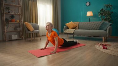 Yaşlı bir kadın oturma odasında yerde bir spor paspasın üzerinde yüzüstü yatıyor. Kadın basının kaslarını geriyor, kollarını uzatıp, vücudunun üst kısmını kaldırıyor.