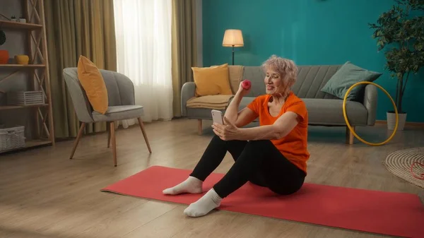一位老年妇女坐在客厅的一个运动垫上 一个女人一只手拿着哑铃 一边练习着她的手 另一边拿着智能手机 一个女人看视频 在视频通话中说话 — 图库照片