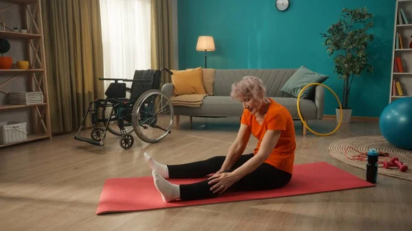 車椅子の横のリビングルームには 身体障害のあるお年寄りの女性が腰を下ろしている 白髪の女性が足のマッサージをします 身体的リハビリテーションの概念 — ストック写真
