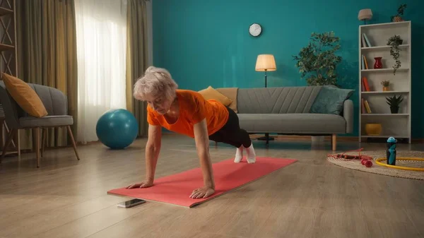 携帯電話の前の床にはスポーツウェアを着た年配の女性が横になっている 女性が腕を伸ばして板運動をする ホームフィットネスコンセプト オンラインスポーツクラス — ストック写真