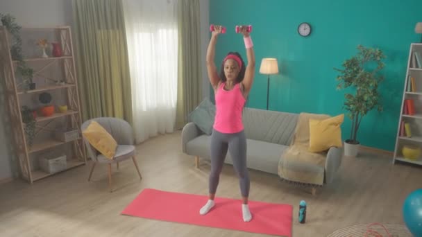 一位非裔美国妇女抬起胳膊 头顶上方的哑铃穿过两边 年轻女子站在客厅的运动垫上锻炼肩膀肌肉 — 图库视频影像
