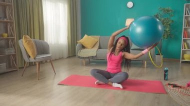 Genç bir kadın spor minderinde bacak bacak üstüne atmış, basın ve kol egzersizi yapıyor. Afrikalı Amerikalı bir kadın kafasının üstünde bir fitness topuyla düz kollarını kaldırır ve yan eğilme hareketi yapar. Ev zindeliği