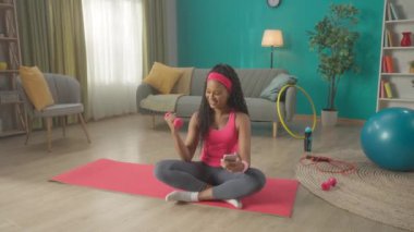 Afrika kökenli Amerikalı bir kadın oturma odasındaki spor paspasının üzerinde bacak bacak üstüne atarak oturuyor. Bir kadın bir elinde dambıl tutarken diğer elinde akıllı telefon, diğer elinde ise elini kullanıyordu. Bir kadın...