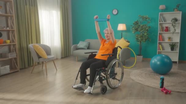 一位坐在轮椅上的老年妇女正在客厅里带着一个运动弹性带进行锻炼 一位白发苍苍的老年妇女锻炼她胳膊和肩膀的肌肉 身体的概念 — 图库视频影像