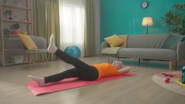 一位老年妇女躺在客厅地板上的一个运动垫上 做运动 这个女人仰卧着 双手反着头 交替地抬起腿 运动的概念 — 图库视频影像