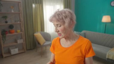 Oturma odasında egzersiz yaptıktan sonra yorgun ve kır saçlı yaşlı bir kadının portresi. Yorgun yaşlı kadın egzersizden sonra su içer. Spor yaşam tarzı, emeklilik, sağlık, sağlık