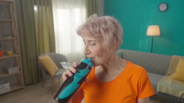 一个疲惫而又满意的白发老妇人在客厅里锻炼后的画像 疲惫的老年妇女运动后喝水 体育生活方式 退休的概念 — 图库视频影像