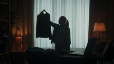 Ne giyeceğine karar veremeyen genç bir kadın. Bir kadın askıda iki bluz tutuyor, omuzlarını silkiyor, hangisini giyeceğine karar veremiyor. Elinde giysileri olan bir kadının karanlık silueti.