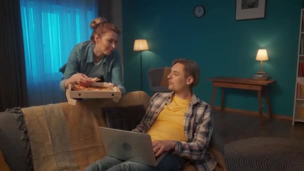 若い男は 彼の膝の上にラップトップでリビングルームでソファの上に横たわっています オンラインゲームをプレイ 彼女の手にピザの箱を持つ女性が彼に来て 彼にスライスを提供し 彼を養う — ストック動画