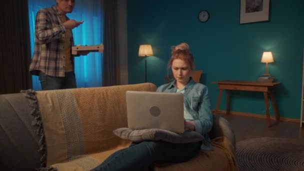若い女性は 彼の膝の上にラップトップでリビングルームでソファの上に横たわっています オンラインゲームをプレイ ピザの箱を手にした男が彼女の所に来て 彼女にスライスを提供し 彼女を養う — ストック動画