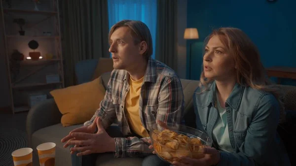 一对夫妇在家里看电视上的体育频道关门了 一名男子和一名女子屏息观看比赛 为他们的球队欢呼 一个女人手里拿着一盘薯片 — 图库照片