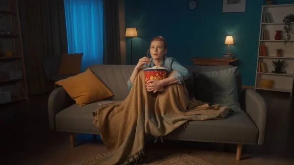恐怖片 在家里看电视上看恐怖片和吃爆米花的年轻女人吓坏了 一个女人拿着毛毯盖住腿 坐在沙发上 手里拿着爆米花 — 图库照片