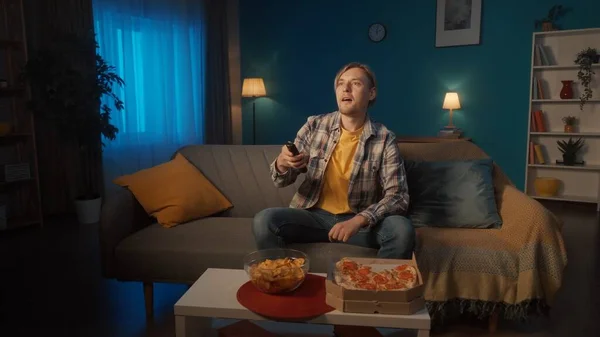 一个男人晚上在家里看电视 一个男人坐在沙发上 用遥控器换频道 有零食 薯片的晚间电视 — 图库照片