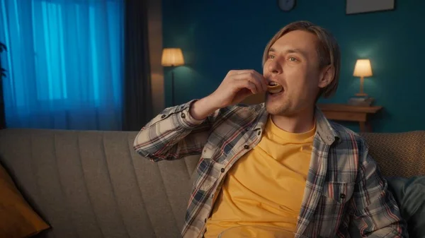 一个男人晚上在家里看电视 一个男人坐在沙发上看电影时吃薯片 沙发上客厅里一个男人的画像靠拢了 — 图库照片