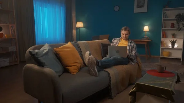 一个年轻人躺在客厅的沙发上 膝盖上放着笔记本电脑 一个面容严肃的人在笔记本电脑上工作或玩耍 看电影 — 图库照片