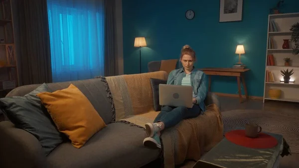 一个年轻的女人躺在客厅的沙发上 膝盖上放着笔记本电脑 一个面容严肃的女人在笔记本电脑上工作或玩耍 看电影 — 图库照片