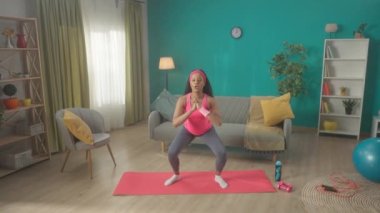 Afrika kökenli Amerikalı bir kadın oturma odasında spor yaparken mekik çekiyor. Bir kadın kalça, kalça, taban ve baldır kaslarını güçlendirir. Kadın ayağa kalkar ve onu sallar.