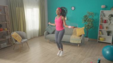 Afro-Amerikalı kadın evde ip atlıyor oturma odasında. Şirin, narin bir kadın evde spor yaparak sağlığına dikkat eder. Spor konsepti, ev fitness, yaşam tarzı. Yavaş çekim