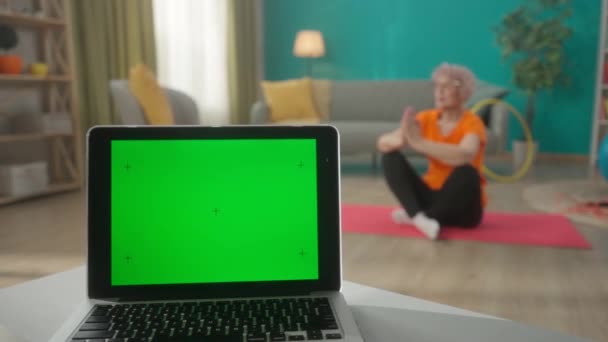 リビングルームのぼやけた背景にテーブルの上に緑の画面を持つノートパソコンが閉じます 背景には スポーツマットの上に蓮の花の位置に座っている高齢者の女性 広告エリア ワークスペース — ストック動画