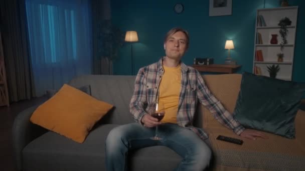 ワインのガラスでテレビの前のソファの上に夜に座って ハード日間の仕事の後に休んでいる 男は赤ワインを飲み 映画を見るのを楽しむ — ストック動画