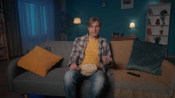 男はポップコーンのプレートとソファに座って 映画を見ています 退屈な表情の男はポップコーンを食べ 携帯電話を拾い ソーシャルネットワーク ビデオを通して見ています 退屈な — ストック動画