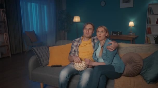 結婚した夫婦は ポップコーンのプレートでソファに抱きつき 映画を見て座っています 男と女がポップコーンを食べながら笑っている 夕方に一緒に映画を見て — ストック動画