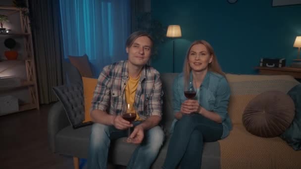 一对年轻夫妇正坐在沙发上 喝着红酒 看电视 一个男人和一个女人正在看一部有兴趣的电影 一个感人的视频 互相看着对方 周末休闲概念 — 图库视频影像