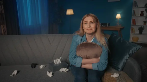 一个多愁善感的女人坐在沙发上 周围是皱巴巴的纸巾 一个女人正在看一部戏剧性的 悲伤的电影或者一部感人的家庭录像 一个悲伤的女人抱着枕头的画像 — 图库照片
