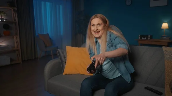 一个女人在晚上坐在家里的沙发上和一个游戏控制器玩一个领事游戏 专心致志的女人按住操纵杆 — 图库照片