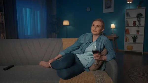 一个年轻的女人正在看电视 喝着红酒 一个女人晚上躺在沙发上 在电视机前休息和放松 — 图库照片